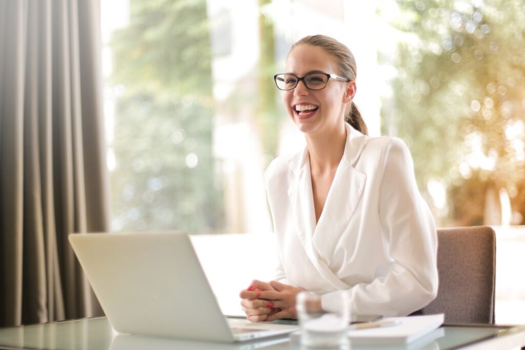 Cheerful female entrepreneur in formal outfit and eyeglasses sitting at table in bright workplace and using netbook while working on project and looking away